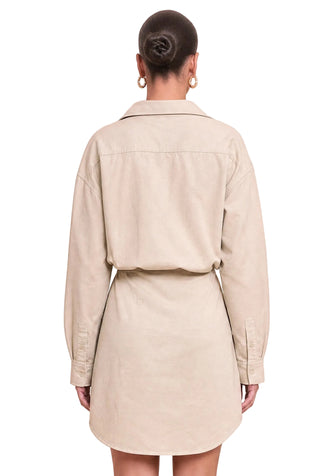 A woman shown from the back wearing a light beige long-sleeve shirt dress with a belted waist, set against a solid pale pink background.