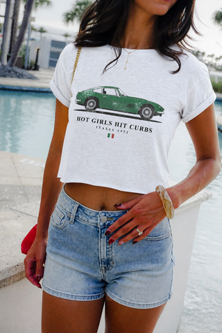 A woman wearing a white cropped graphic t-shirt with a green car print and the text 'HOT GIRLS HIT CURBS' by Piazza 1972, paired with light-wash denim shorts, standing by a swimming pool.