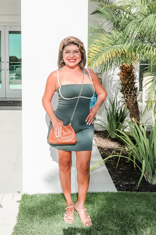 A woman stands outdoors wearing a fitted sage green strapless mini dress with white trim, holding a brown leather clutch, and wearing clear high-heel sandals. The background features a white wall, a palm tree, and green grass.