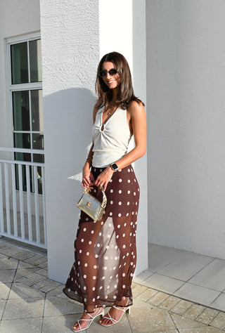 A woman stands outdoors in a white stucco building setting, wearing a sleeveless cream top and a brown sheer maxi skirt with white polka dots. She is holding a small metallic handbag and wearing strappy high-heeled sandals.