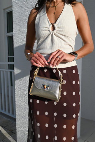 A woman wearing a cream ribbed halter top and a brown sheer skirt with white polka dots, holding a metallic silver handbag with gold hardware.
