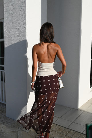 A woman is shown from behind wearing a brown strapless maxi skirt with white polka dots over a white top, standing outdoors against a white stucco wall.