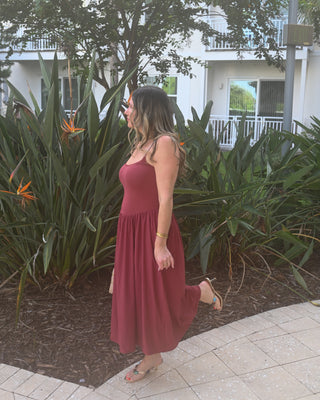 A woman wearing a sleeveless rust-red midi dress walks outdoors on a paved path with tropical plants and a white building in the background.