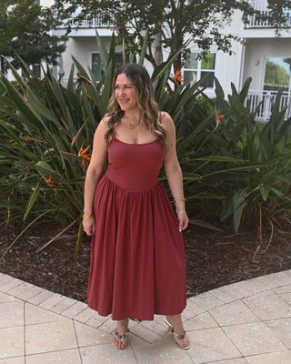 A woman wearing a sleeveless rust-red midi dress with a gathered waist and flared skirt, standing outdoors on a tiled patio in front of tropical plants and a white building.