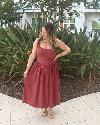 A woman wearing a sleeveless rust-colored midi dress with a gathered waist and flared skirt, standing outdoors on a tiled patio in front of tropical plants and a white house.