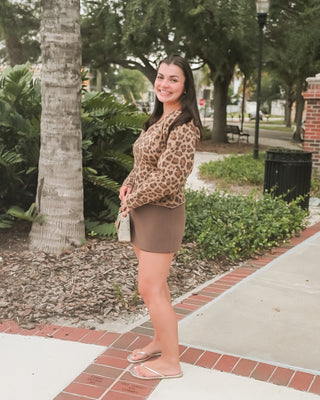 A woman posing outdoors on a brick sidewalk next to a palm tree, wearing a leopard print long-sleeve top and a brown mini skirt.