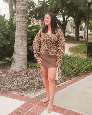 A woman stands outdoors on a brick sidewalk wearing a brown and beige leopard-print long-sleeve crop top with a matching brown mini skirt, holding a small woven handbag.