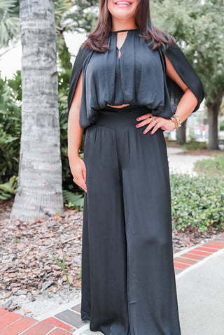 A woman wearing a black two-piece outfit consisting of a short-sleeve keyhole top with draped cap sleeves and matching wide-leg pants, standing outdoors on a sidewalk with palm trees and greenery in the background.