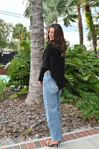 A woman wearing a black long-sleeve knit top and light-wash wide-leg jeans stands outdoors next to a palm tree, looking back over her shoulder.