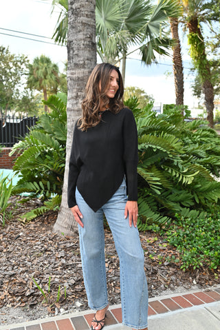 A woman wearing a black long-sleeve asymmetrical top and light-wash wide-leg jeans stands outdoors in front of lush green foliage and palm trees.