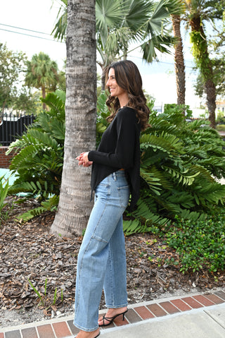 A woman stands outdoors in a tropical garden setting, wearing a black long-sleeve top, light-wash wide-leg blue jeans, and black strappy heels.