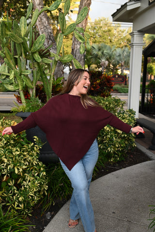 A woman wearing a loose-fitting, oversized burgundy knit sweater and light blue jeans stands outdoors on a sidewalk with her arms outstretched, surrounded by green plants and trees.