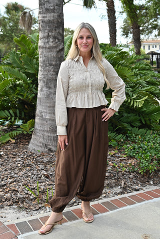 A woman stands outdoors in front of palm trees and greenery, wearing a cream ribbed long-sleeve peplum-style blouse with a smocked chest panel and dark brown loose harem-style pants.