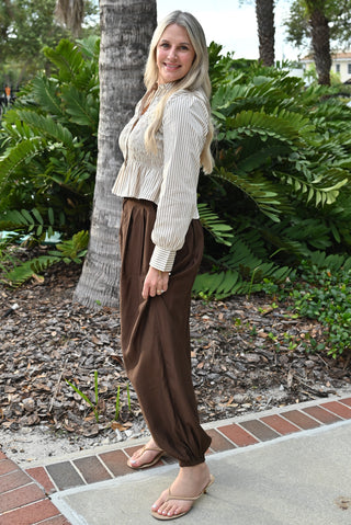 A woman stands outdoors on a brick walkway wearing a cream ribbed long-sleeve cropped top and brown wide-leg pants, with green foliage and palm trees in the background.