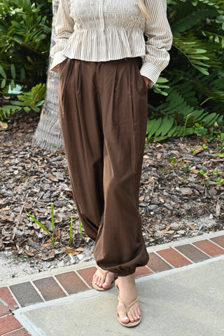A person wearing loose brown wide-leg pants and a cream striped long-sleeve top, standing outdoors on a brick walkway with mulch and green plants in the background.