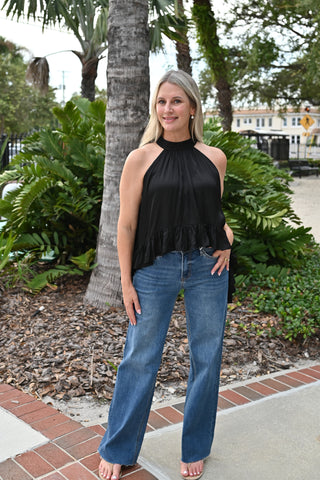 A woman stands outdoors in front of palm trees and greenery, wearing a sleeveless black halter top with a ruffled hem and blue wide-leg jeans.