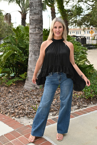 A woman wearing a black halter-neck ruffled top and blue wide-leg jeans, standing outdoors on a brick walkway with palm trees and greenery in the background.