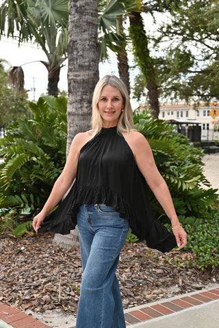 A woman wearing a black halter-neck ruffled top and blue jeans stands outdoors in a landscaped area with palm trees and greenery.