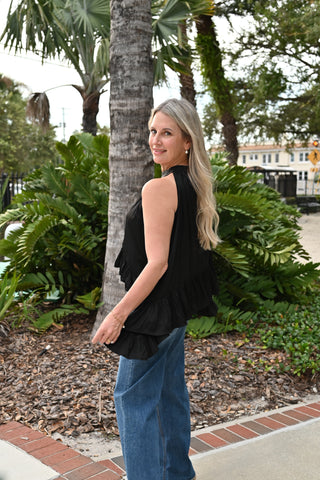 A woman with blonde hair stands outdoors in front of a palm tree, wearing a sleeveless black ruffled top and blue jeans.