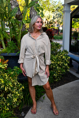 A woman with gray hair models a light beige, long-sleeve shirt dress with a tie waist, standing outdoors on a concrete walkway surrounded by green plants and a house porch.