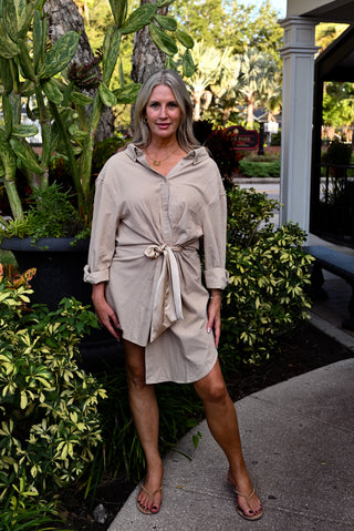 A woman wearing a beige button-down shirt dress tied at the waist, standing outdoors on a concrete walkway surrounded by green plants and trees.