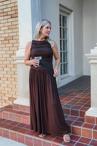 A woman wearing a sleeveless, high-neck, floor-length brown maxi dress stands on red brick steps outside a building with beige columns and brick walls.