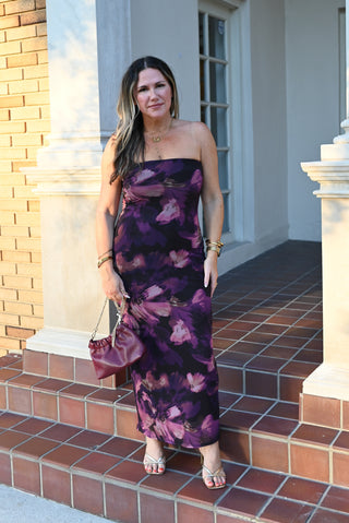 A woman stands on outdoor brick steps in front of a building, wearing a strapless purple floral maxi dress with a draped bodice, holding a matching purple handbag.