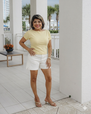 Woman in a yellow top and white shorts standing on a patio with palm trees in the background.