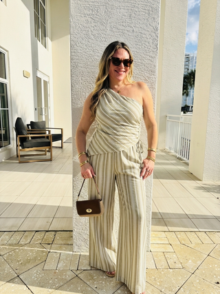 A woman stands on a sunlit outdoor patio wearing a beige and cream vertically striped one-shoulder jumpsuit. She is holding a small brown handbag and wearing sunglasses, with a modern white building and tiled floor in the background.