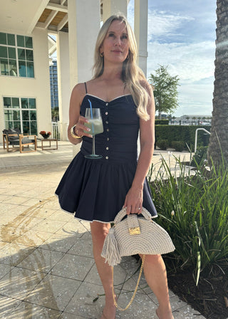 Woman in a navy dress holding a drink and a straw bag outdoors.