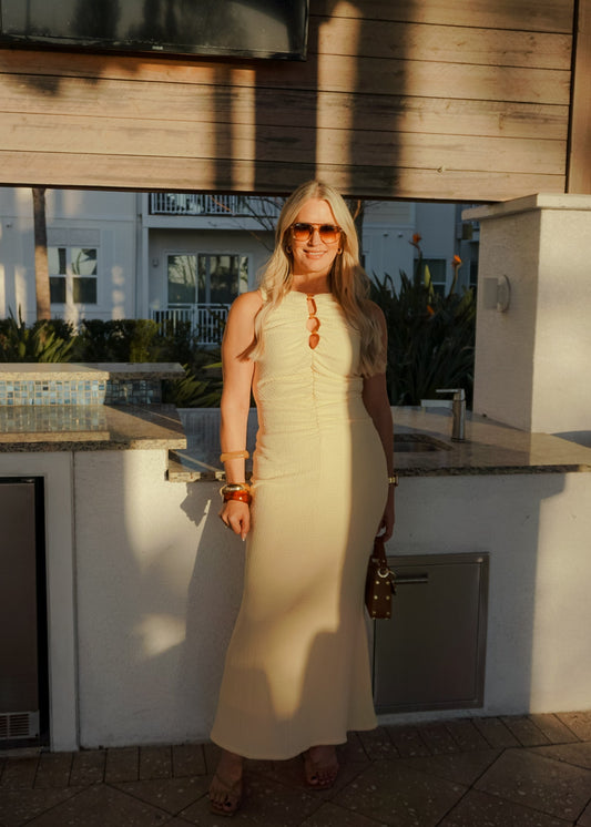 Woman in a beige dress standing outdoors near a wooden wall.