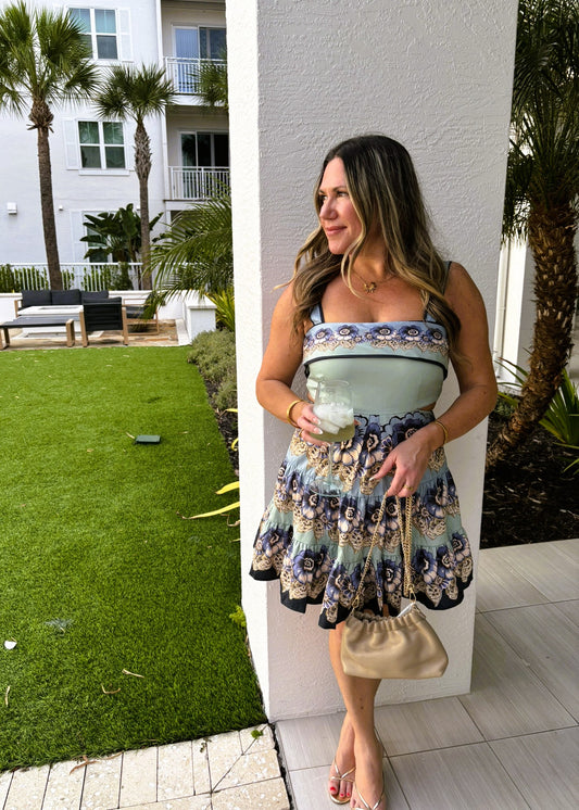 Woman in a floral dress holding a light blue handbag, standing against a white wall with palm trees and a building in the background.