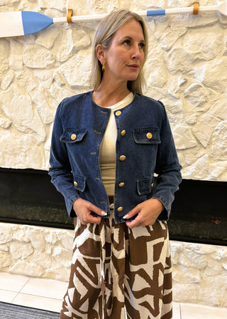 Woman wearing a denim jacket and patterned pants standing against a stone wall.