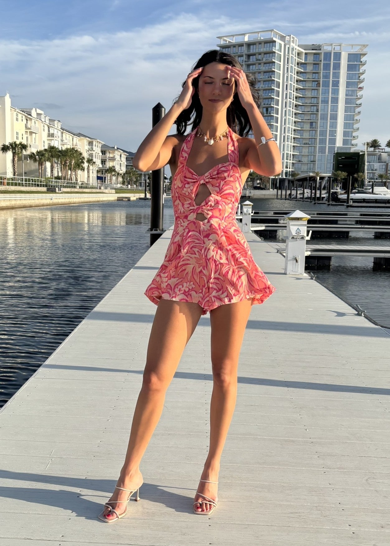 Woman in a pink floral dress standing on a dock with buildings and water in the background