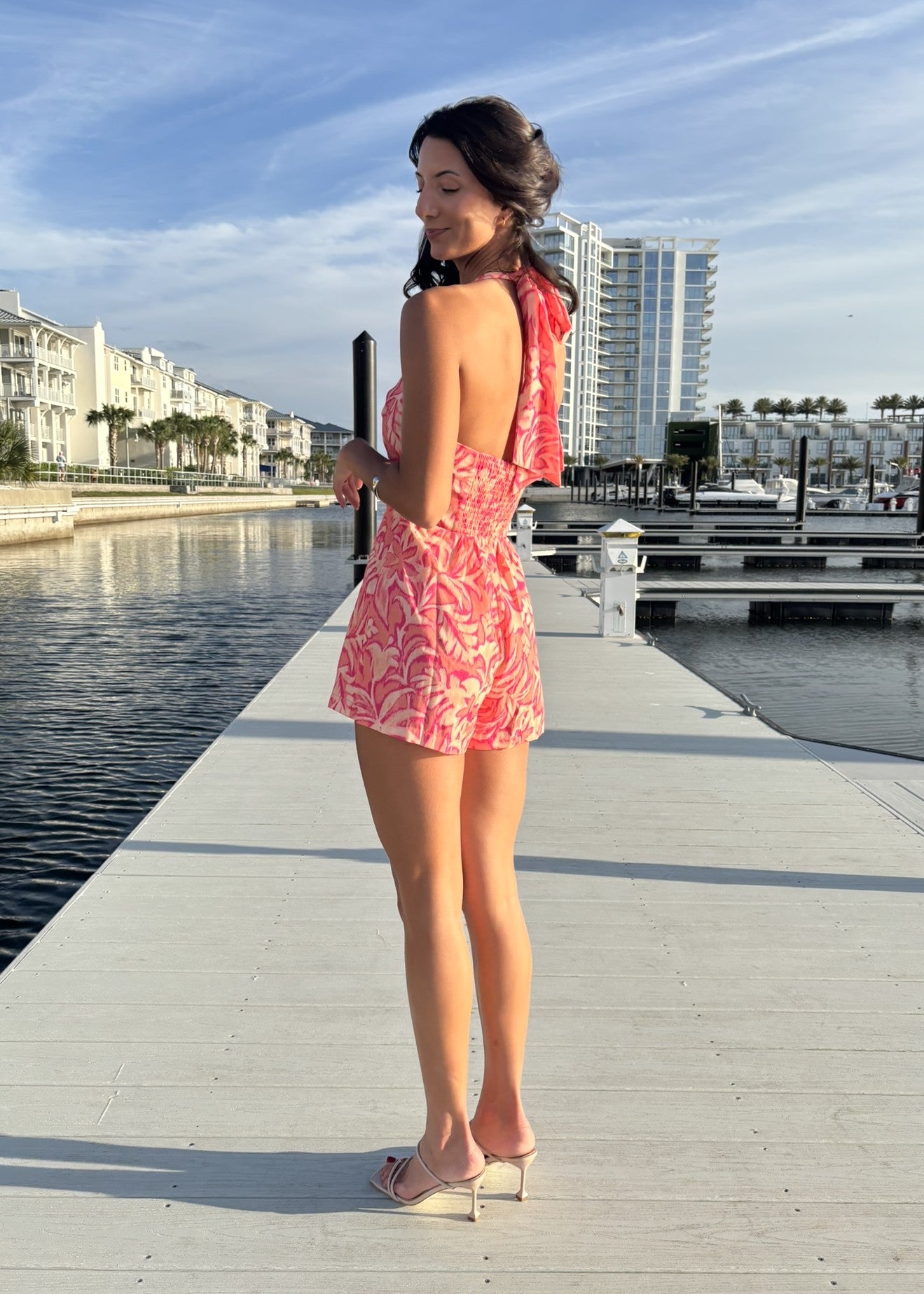 Woman in a pink floral dress standing on a dock by a waterfront with buildings in the background.