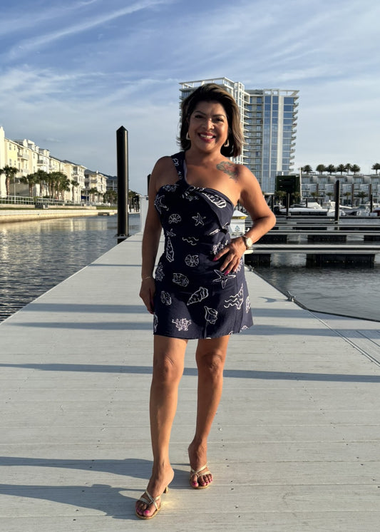 Woman in a navy dress with white patterns standing on a dock by a waterfront with buildings in the background.
