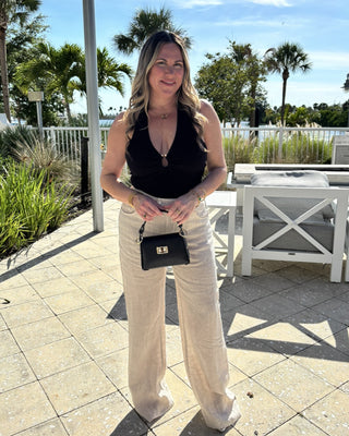 Woman in black top and beige pants standing outdoors with palm trees and a clear sky in the background.