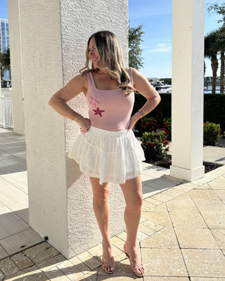 Woman in a pink tank top and white skirt standing outdoors near a building.