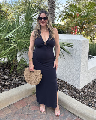 Woman in a black dress holding a wicker handbag in an outdoor setting with plants and a white wall.