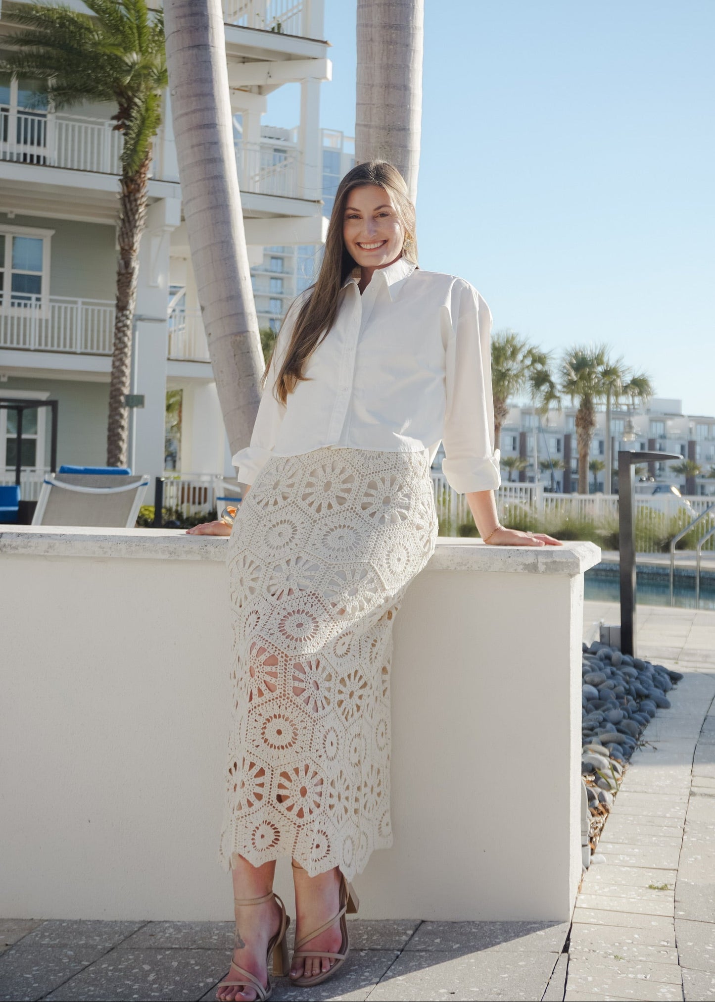 Woman in a white blouse and lace skirt standing on a balcony with palm trees and buildings in the background.
