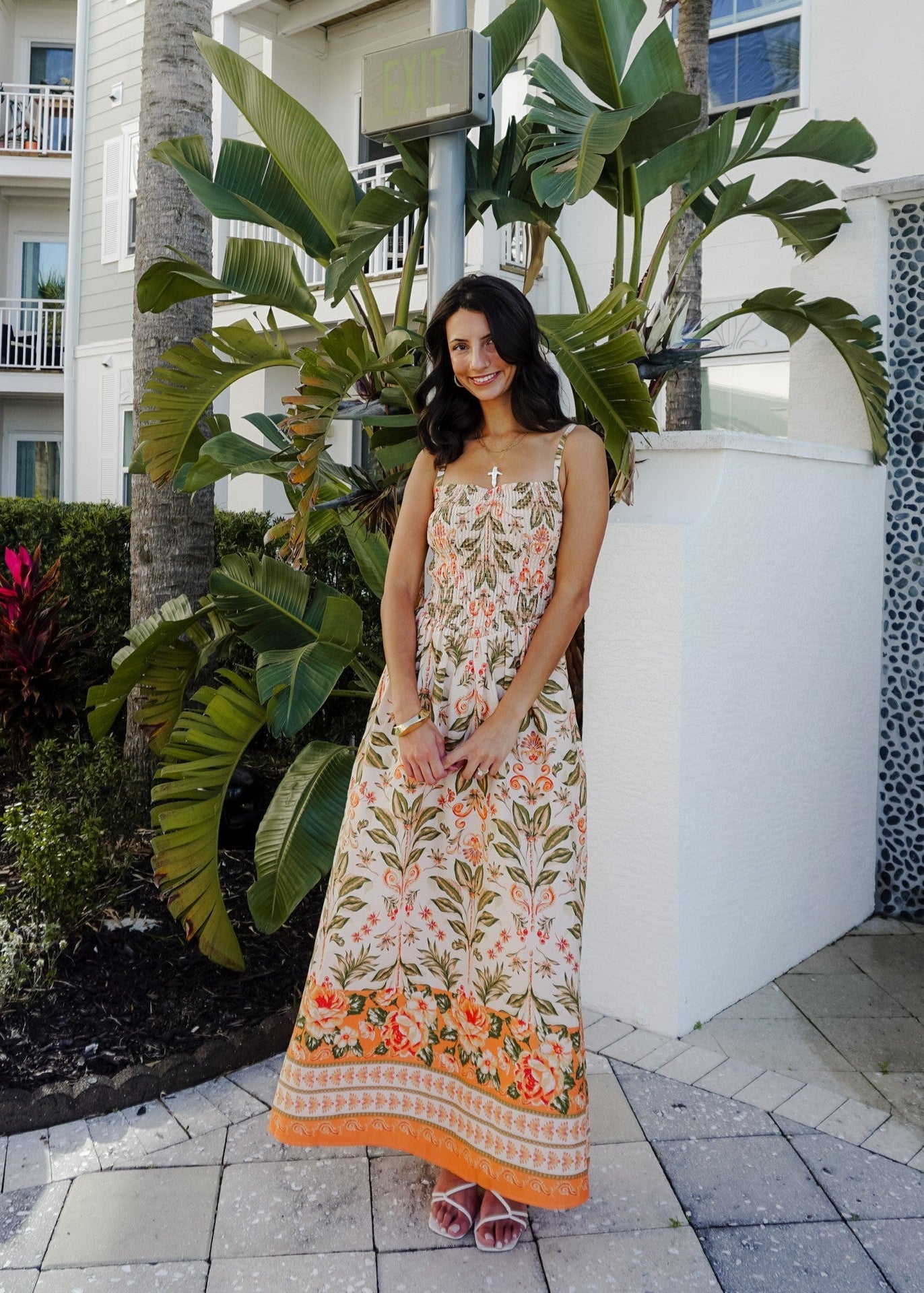 Woman in a floral dress standing outdoors near plants and a building.