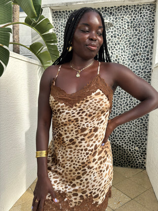 Woman wearing a leopard print dress standing outdoors with a patterned wall and plant in the background.