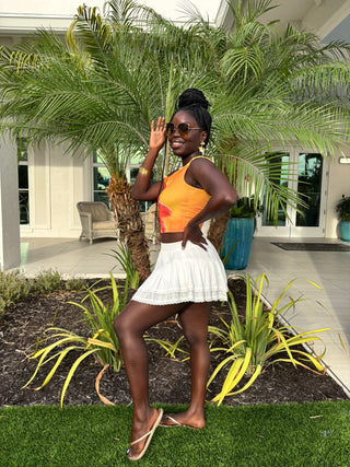 Woman in an orange top and white skirt sitting on a stone wall with palm trees and a building in the background.