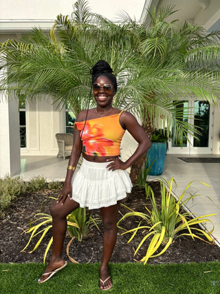 Woman in colorful top and white skirt standing in a garden with palm trees and a house in the background.
