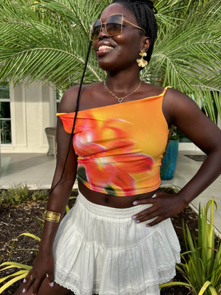 Woman wearing an orange floral top and white skirt outdoors with greenery in the background