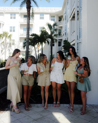 Group of women standing together on a sunlit street with palm trees and a building in the background.