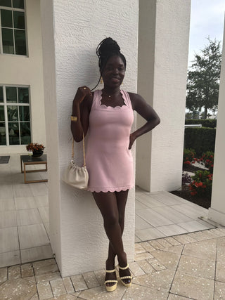 Woman in a pink dress standing against a white brick wall outdoors.