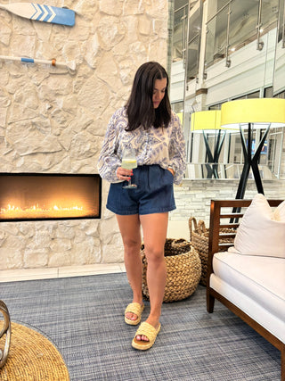 A woman stands in a modern indoor setting with a stone fireplace, wearing a light blue and white patterned long-sleeve top, dark blue denim shorts, and pale yellow slide sandals. She is holding a glass of white wine.
