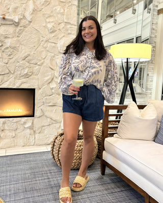 A woman stands in a modern living room wearing a light beige and gray patterned long-sleeve blouse, dark blue denim shorts, and beige slide sandals, holding a glass of white wine.