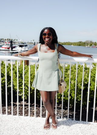Woman in a light green dress standing on a dock with boats and greenery in the background.